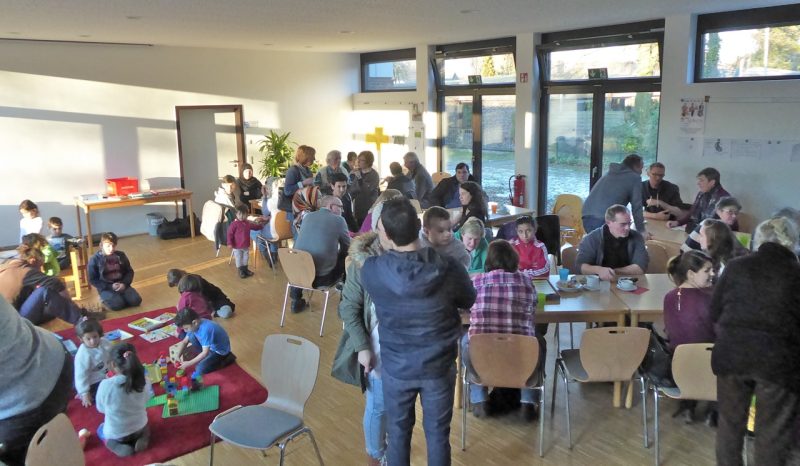 Viele Erwachsene sitzen und stehen an Tischen im Gemeindesaal, Kinder spielen auf einem Teppich